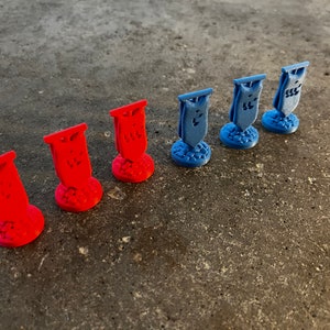 K&ouml;nnte beinhalten: Sieben Miniaturflaggen, drei rote und vier blaue, stehen auf einer grauen Oberfl&auml;che. Jede Flagge hat ein einzigartiges Design und ist auf einem kleinen Sockel montiert.