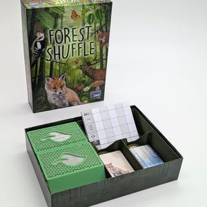 May include: A board game box titled "Forest Shuffle" with illustrations of forest animals, including a fox, woodpecker, and squirrel. The game box is next to an open box containing green card holders, cards, and a game board.
