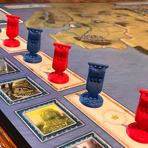 K&ouml;nnte beinhalten: Ein Spielbrett mit roten und blauen Flaggen darauf. Die Flaggen haben das Wort "The" darauf. Das Brett hat einen blauen Hintergrund und eine Karte einer Landmasse darauf. Das Brett hat auch ein paar verschiedene Felder mit Namen darauf, wie "Woodland Realm" und "Lorien".