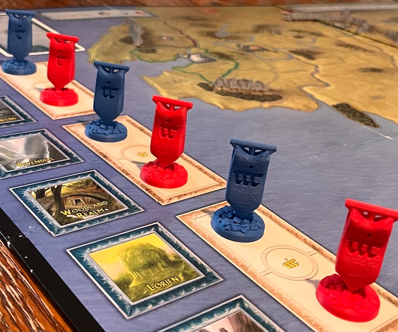 K&ouml;nnte beinhalten: Ein Spielbrett mit roten und blauen Flaggen darauf. Die Flaggen haben das Wort "The" darauf. Das Brett hat einen blauen Hintergrund und eine Karte einer Landmasse darauf. Das Brett hat auch ein paar verschiedene Felder mit Namen darauf, wie "Woodland Realm" und "Lorien".