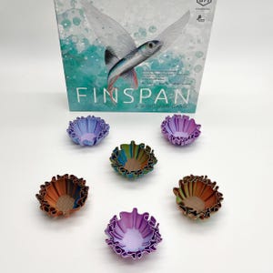 May include: The board game "Wingspan" is displayed with six colorful, ruffled-edge bowls. The bowls are in shades of purple, brown, green, and orange. The game box features a flying fish illustration and the game title.