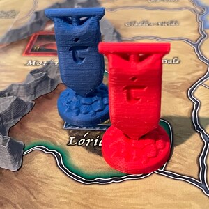 K&ouml;nnte beinhalten: Zwei 3D-gedruckte Spielfiguren, eine blaue und eine rote, mit einem stilisierten Buchstaben 'T' auf jeder. Die Figuren befinden sich auf einem Spielbrett mit braunem Hintergrund.