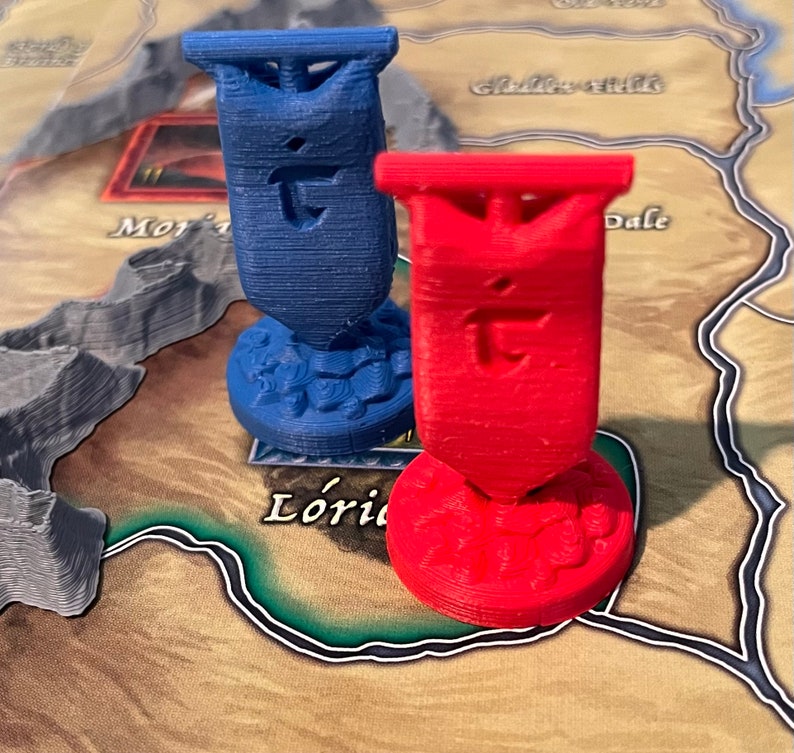 K&ouml;nnte beinhalten: Zwei 3D-gedruckte Spielfiguren, eine blaue und eine rote, mit einem stilisierten Buchstaben 'T' auf jeder. Die Figuren befinden sich auf einem Spielbrett mit braunem Hintergrund.