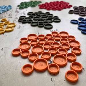 Puede incluir: Primer plano de un conjunto de fichas de juego de color naranja con la palabra "Play" inscrita en cada ficha. Las fichas est&aacute;n dispuestas en semic&iacute;rculo sobre una superficie de madera blanca.