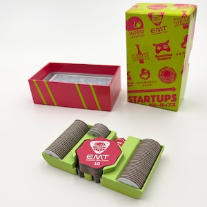 May include: A green box with the text "Startups" and various animal logos in pink. The box is open and shows a red card with the text "EMT Elephant Mars Travel 10". The card is in a green holder with two stacks of brown cardboard circles.