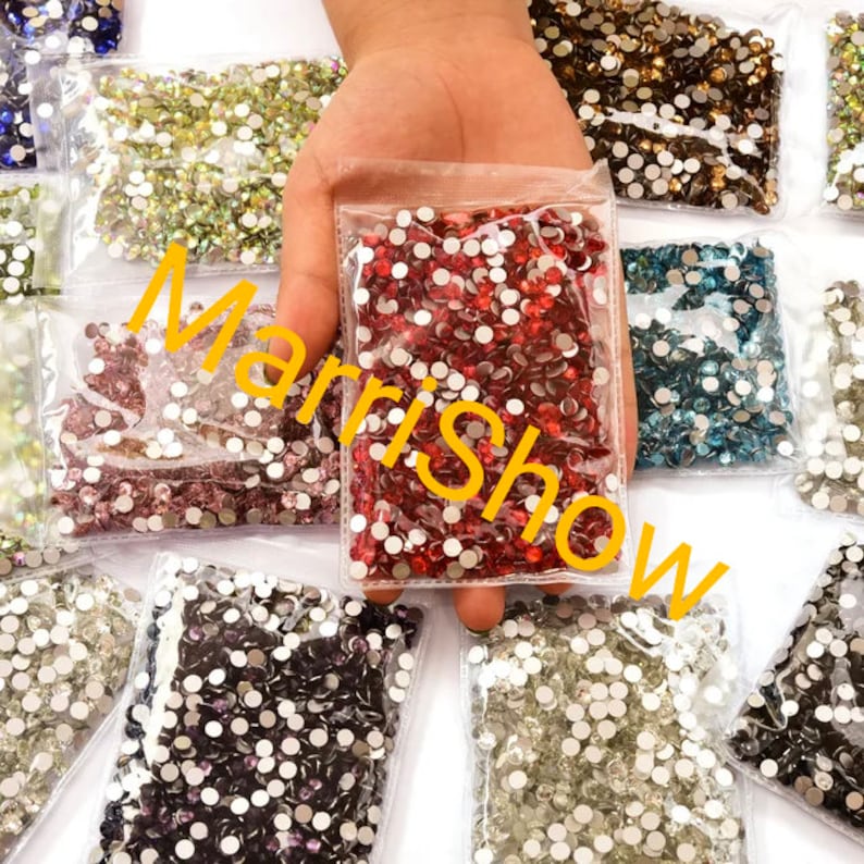 May include: A hand holding a clear plastic bag filled with red rhinestones, surrounded by other bags of various colored rhinestones. The text "Marrishow" is overlaid on the image in yellow.