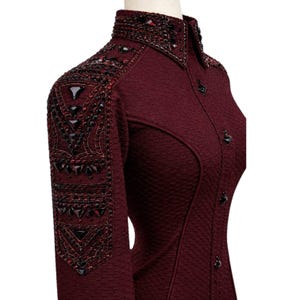 May include: A deep burgundy long-sleeved shirt featuring a textured fabric, a high collar, and a button-down front. The shoulders and collar are heavily embellished with dark red and black triangular crystals and intricate beadwork patterns.
