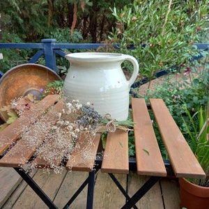 May include: A white ceramic jug with a speckled design sits on a wooden table with dried flowers. The jug has a handle and a spout.