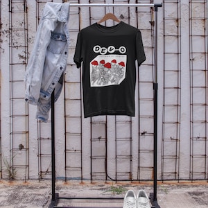 Op de afbeelding: Zwart T-shirt met een witte afbeelding van een band genaamd DEVO. De afbeelding toont een groep mensen met rode hoeden en de tekst "Freedom of Choice".