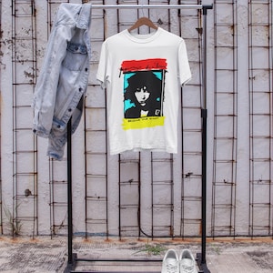 Pode incluir: Camiseta branca com uma imagem em preto e branco de uma pessoa com acentos azuis e amarelos. O texto "Because the night" é impresso abaixo da imagem.