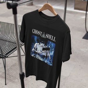 May include: Black t-shirt featuring a graphic print of a woman holding a gun. The text "Ghost in the Shell" is printed in white above the graphic.