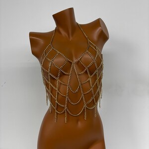 May include: A gold chainmail body chain with a delicate, intricate design. The chainmail is made of small, linked metal rings, creating a web-like pattern. The chainmail is designed to be worn as a top, with a halter neckline and a flowing, fringed bottom.