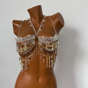 May include: A beaded bra top with a gold and clear crystal design. The bra top has a halter style neckline and features a fringe detail.