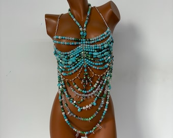 May include: A turquoise beaded body chain with gold-colored chain straps. The body jewelry features various sizes of turquoise beads, clear crystals, and small white beads. The design is displayed on a mannequin.