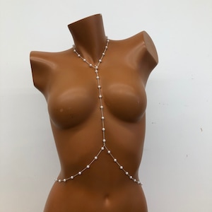 May include: A silver body chain with white pearl beads. The chain is long and drapes down the front of the body.