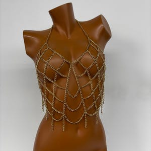 May include: Gold chain body jewelry with a delicate, intricate design. The chain is made of small, linked rings that form a web-like pattern. The jewelry is designed to be worn as a top or a statement piece.