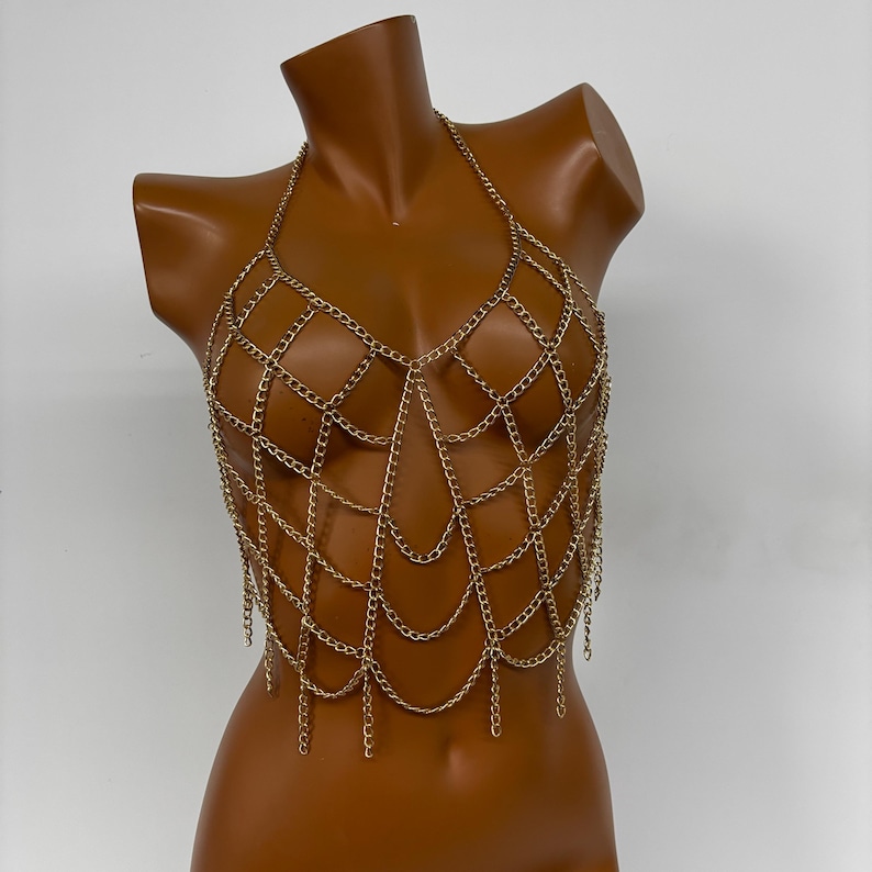 May include: Gold chain body jewelry with a delicate, intricate design. The chain is made of small, linked rings that form a web-like pattern. The jewelry is designed to be worn as a top or a statement piece.