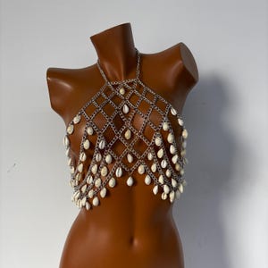 Puede incluir: Un top de malla de plata con conchas blancas colgando de él. El top está en un maniquí.