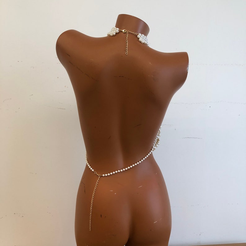 May include: A white pearl body chain with a gold chain connecting the necklace and waist chain. The chain is made of small, round pearls.