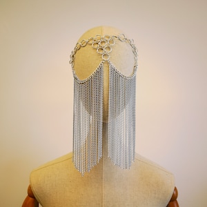 Puede incluir: Un tocado de cadena plateada con cadenas largas y colgantes. El tocado está hecho de círculos entrelazados y se lleva en un maniquí.