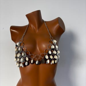 Puede incluir: Top halter plateado con cuello de cadena y detalles circulares. El top está adornado con conchas blancas y discos plateados, creando un efecto en capas. El top se exhibe en un maniquí.