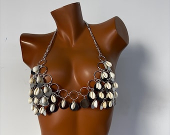 May include: A silver-toned halter top with a chain neckline and circular accents. The top is adorned with white seashells and silver-colored discs, creating a layered, decorative effect. The top is displayed on a mannequin.