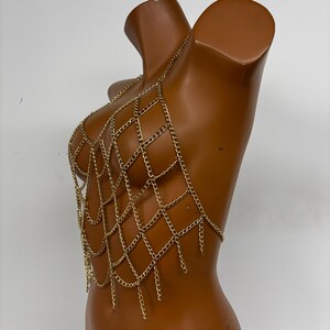May include: A gold chain body jewelry piece with a geometric design. The chain is made of multiple layers of small linked chains that form a web-like pattern.