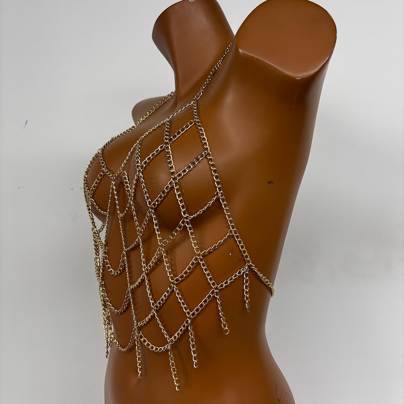 May include: A gold chain body jewelry piece with a geometric design. The chain is made of multiple layers of small linked chains that form a web-like pattern.
