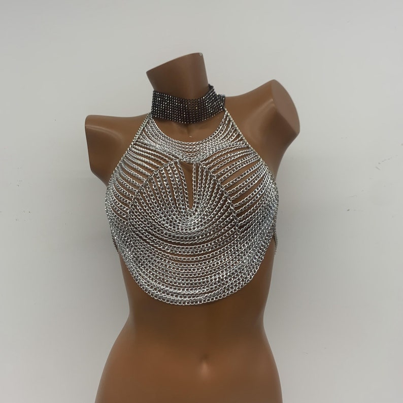 May include: A silver chainmail halter top with a black choker necklace. The top is made of interlocking metal rings and has a V-shaped neckline.