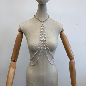 May include: A white pearl body chain with a gold chain at the top. The chain is layered and has multiple strands of pearls.