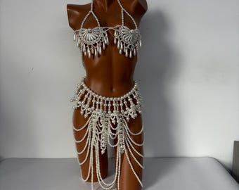 May include: A white pearl bikini set on a brown mannequin. The top features a halter neck and decorative sunburst-shaped accents. The bottom is a skirt with multiple strands of pearls.