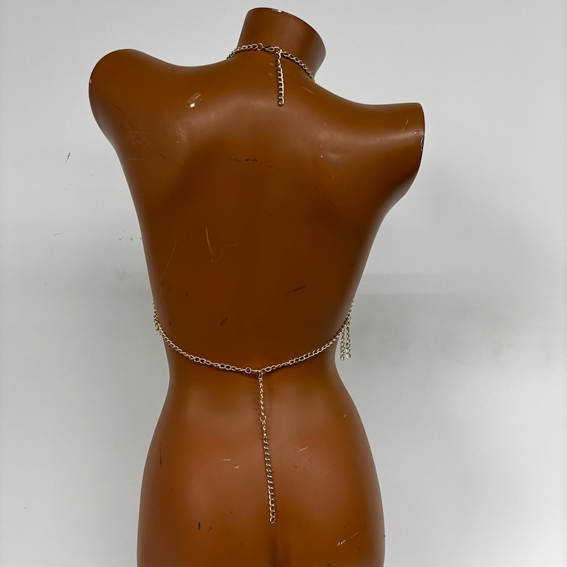 May include: Gold chain body jewelry with a delicate chain and dangling charms. The jewelry is displayed on a brown mannequin.