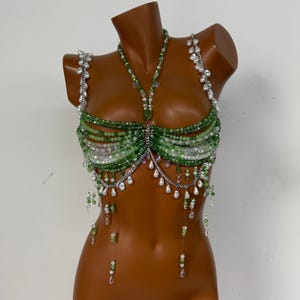 Può includere: Top verde con perline con dettagli a catena argentata e perline pendenti a forma di perla. Il top è esposto su un manichino e presenta un design all'americana con più file di perline verdi e un design centrale con perline.