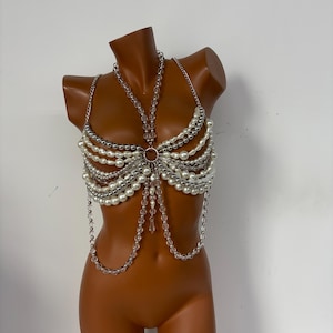 Puede incluir: Un bustier hecho de perlas blancas de imitación, cuentas plateadas y cuentas transparentes con forma de cristal. La parte superior presenta un anillo metálico circular en el centro y correas de cadena. El bustier se exhibe en un maniquí.