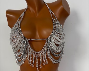May include: A silver-toned halter top with clear and silver beads. The top features a chain neckline and a beaded fringe that hangs down. The beads are arranged in rows and clusters, creating a shimmering effect. The top is designed to be worn as a statement piece.