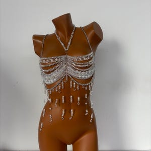 May include: A white beaded bra top with a delicate chain design and dangling beads. The top is made of clear plastic beads and has a silver chain.