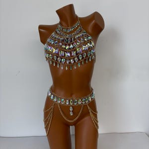 May include: A gold-toned body chain set with iridescent square and teardrop-shaped crystals. The set includes a halter top, a waist chain, and a chain that drapes between the legs. The crystals reflect multiple colors.