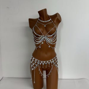 May include: A silver body chain with crystal embellishments. The chain features a layered design with multiple rows of crystals and a dangling pendant. The chain is worn over a nude-colored mannequin.