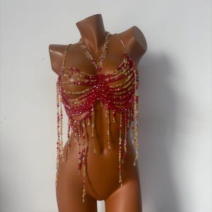 May include: A red and gold beaded top with a matching necklace. The top features a halter style with rows of beads and dangling strands. The beads are a mix of red and gold, creating a shimmering effect. The necklace has a similar design.