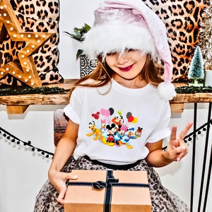 May include: A young girl wearing a pink and white Santa hat holds a wrapped gift box. She is wearing a white t-shirt with a colorful print of Disney characters and balloons. The girl is sitting on a leopard print chair.