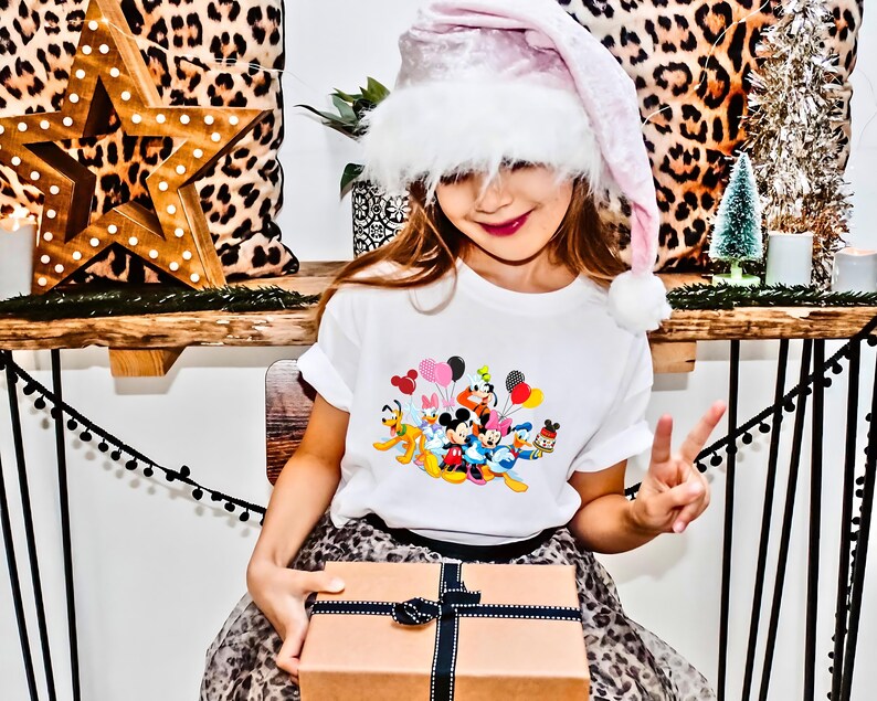 May include: A young girl wearing a pink and white Santa hat holds a wrapped gift box. She is wearing a white t-shirt with a colorful print of Disney characters and balloons. The girl is sitting on a leopard print chair.