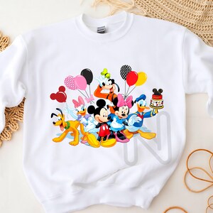 May include: White sweatshirt with a colorful graphic print of Mickey Mouse and friends celebrating with balloons and a birthday cake.