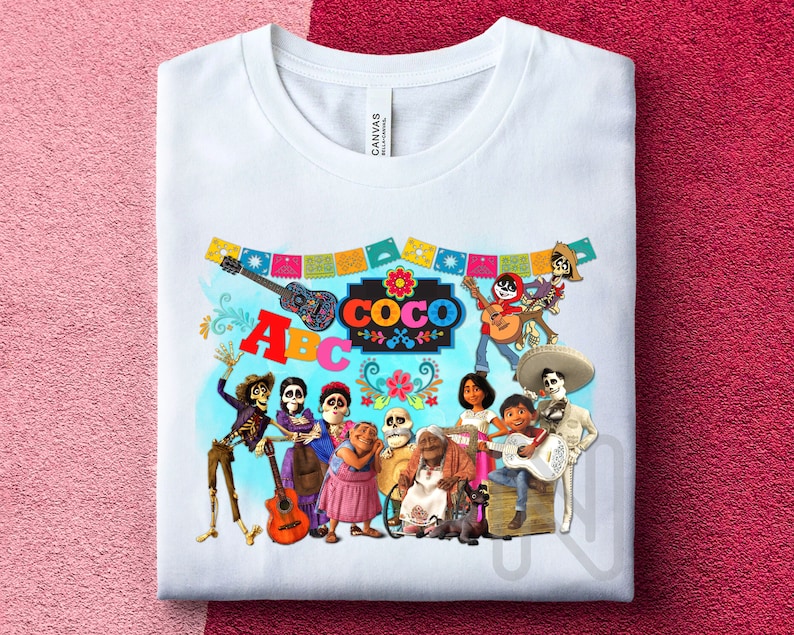 May include: White t-shirt with a colorful graphic of characters from the movie Coco. The graphic includes the characters playing musical instruments and the word "Coco" in colorful letters. The graphic also includes a banner with the letters "ABC".