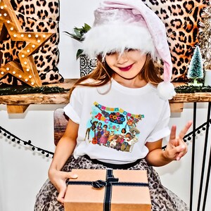 May include: A young girl wearing a white t-shirt with a colorful "Coco" graphic and a pink Santa hat holds a wrapped gift box. The girl is sitting on a leopard print chair in front of a wooden table with a star-shaped ornament and a small Christmas tree.