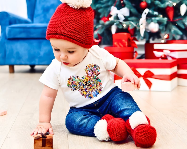 May include: A toddler wearing a red knit hat and a white t-shirt with a colorful Mickey Mouse design. The toddler is wearing blue jeans and red and white knitted booties. The toddler is sitting on a wooden floor in front of a Christmas tree.