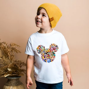 May include: A white t-shirt with a colorful Mickey Mouse head design made up of many Disney characters. The shirt is being worn by a young child wearing a yellow beanie.