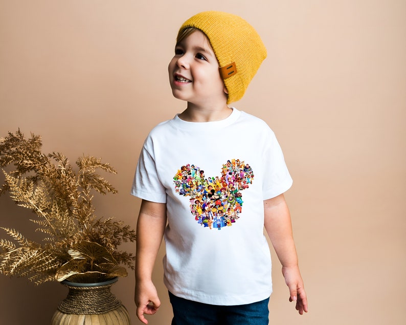 May include: A white t-shirt with a colorful Mickey Mouse head design made up of many Disney characters. The shirt is being worn by a young child wearing a yellow beanie.