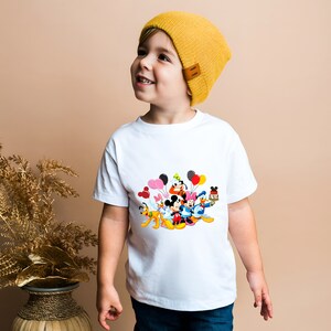 May include: A white t-shirt with a colorful graphic of Mickey Mouse and friends holding balloons. A young child is wearing the shirt and a yellow beanie.