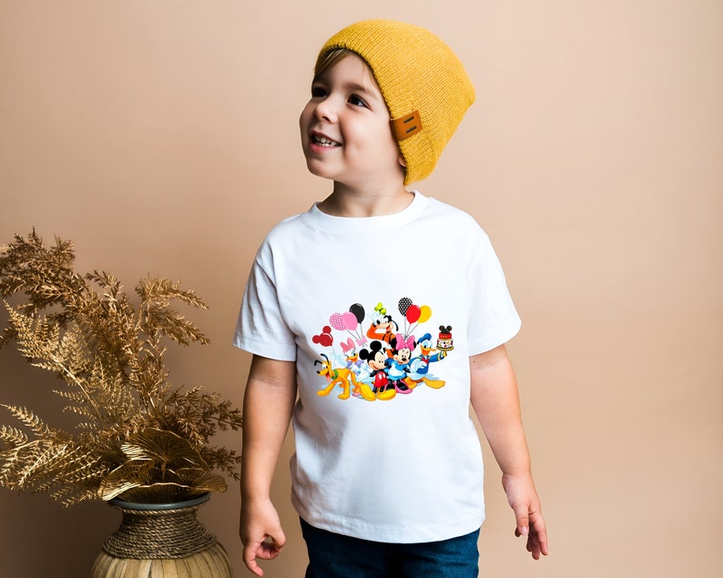May include: A white t-shirt with a colorful graphic of Mickey Mouse and friends holding balloons. A young child is wearing the shirt and a yellow beanie.