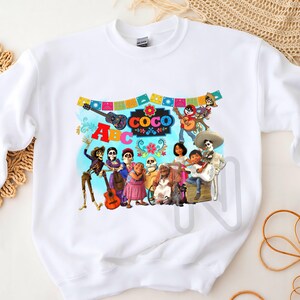 May include: White sweatshirt with a colorful graphic print of characters from the movie Coco. The print includes the characters Miguel, Hector, Dante, and others. The text "ABC COCO" is also included in the print.